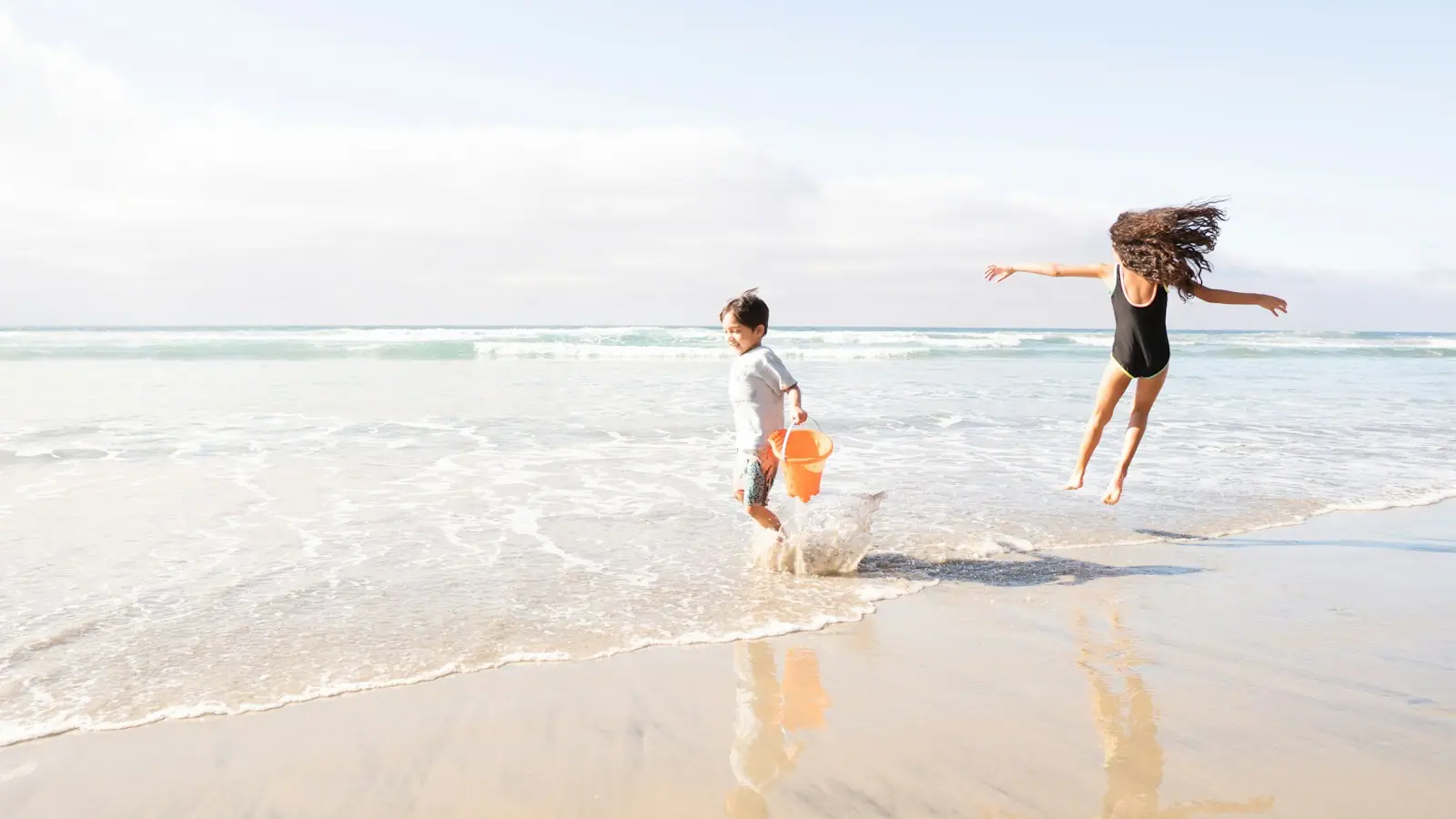 Two children playing in shallow water at the edge of a pale, flat beach