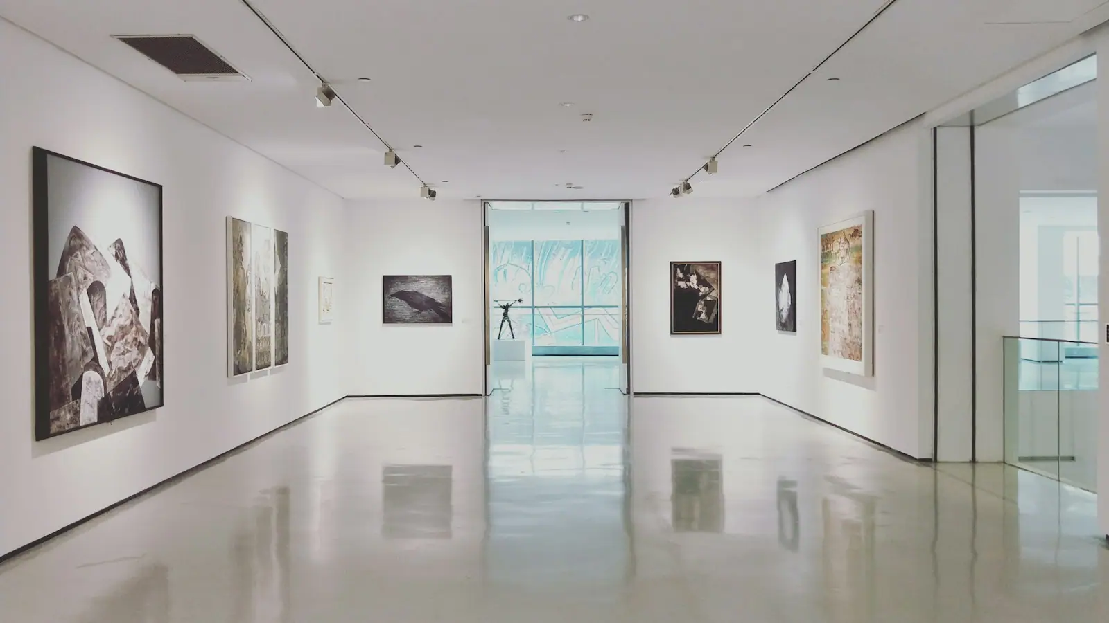 A bright, white-walled gallery with framed works hung along both sides and daylight at the far end