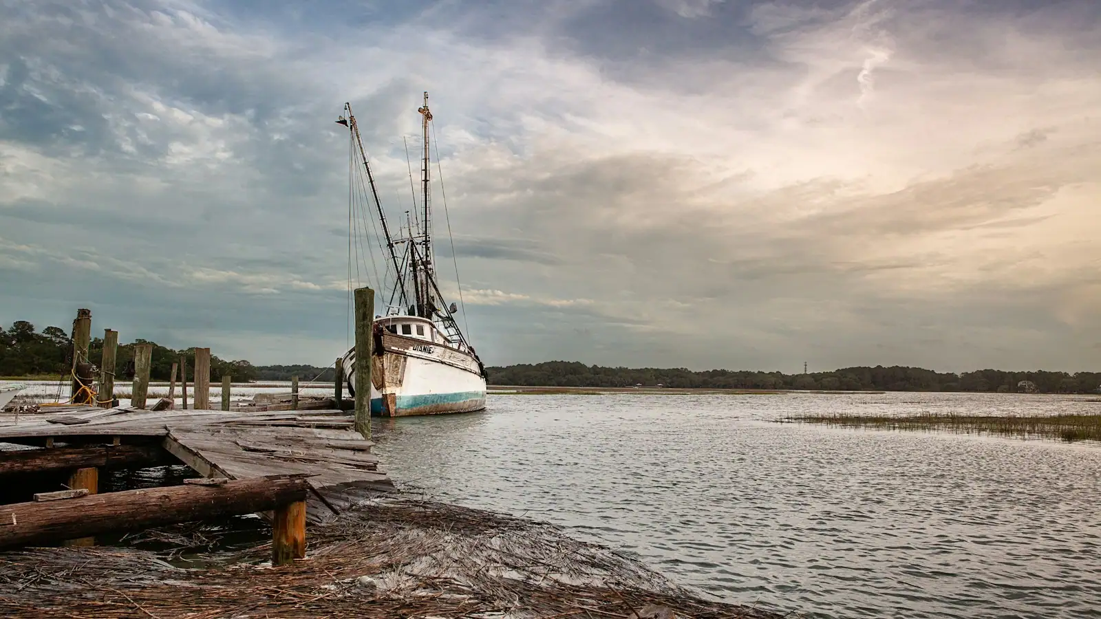 An older fishing boat tied to a weathered timber dock on a back-bay channel under a wide evening sky